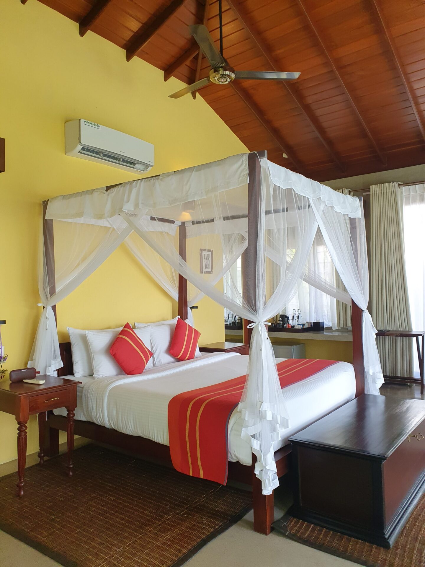 Canopy bed in rich wood with crisp white linens, colourful cushions and a warm yellow accent wall at Aarunya
