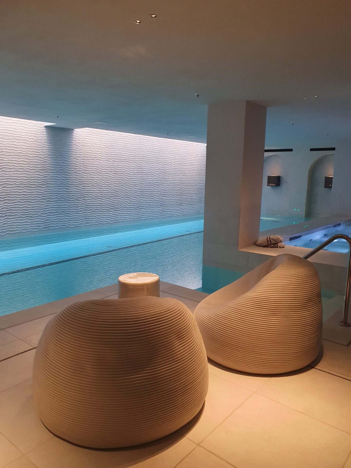 Elegant spa at Rosewood Amsterdam featuring round relaxation chairs in the foreground and a serene indoor pool in the background