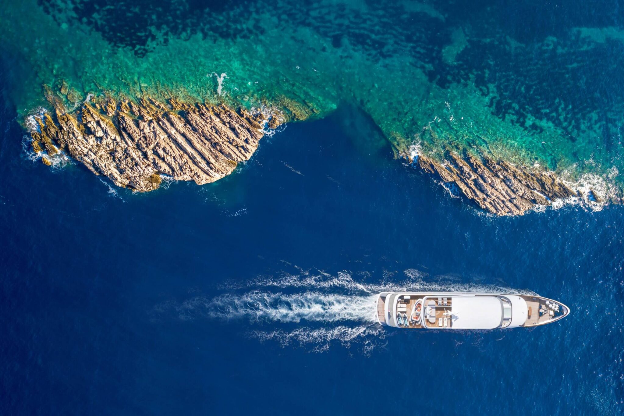Aerial view of a luxury yacht sailing between rocky islets in the Adriatic Sea, near the Croatian coast, surrounded by deep blue and turquoise waters