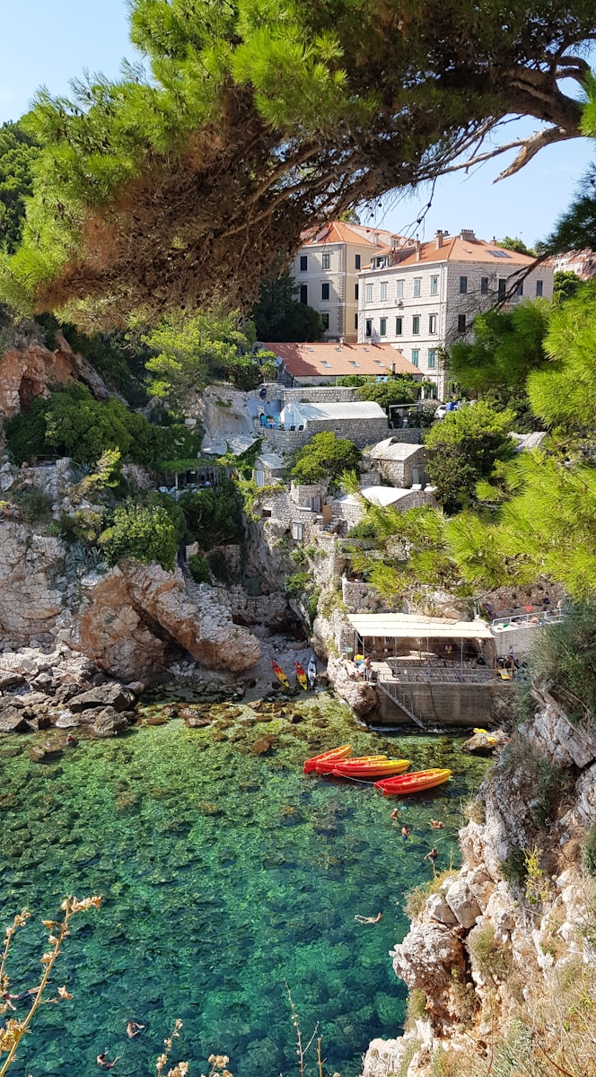 Secluded swimming cove near Dubrovnik, with stone houses built into the rocky cliffside and bright orange and yellow kayaks floating along the shore.